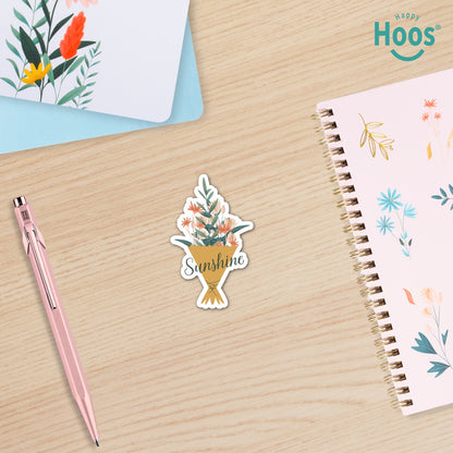 Sticker with floral design and 'Sunshine' text on a wooden surface with a pen and notebook.