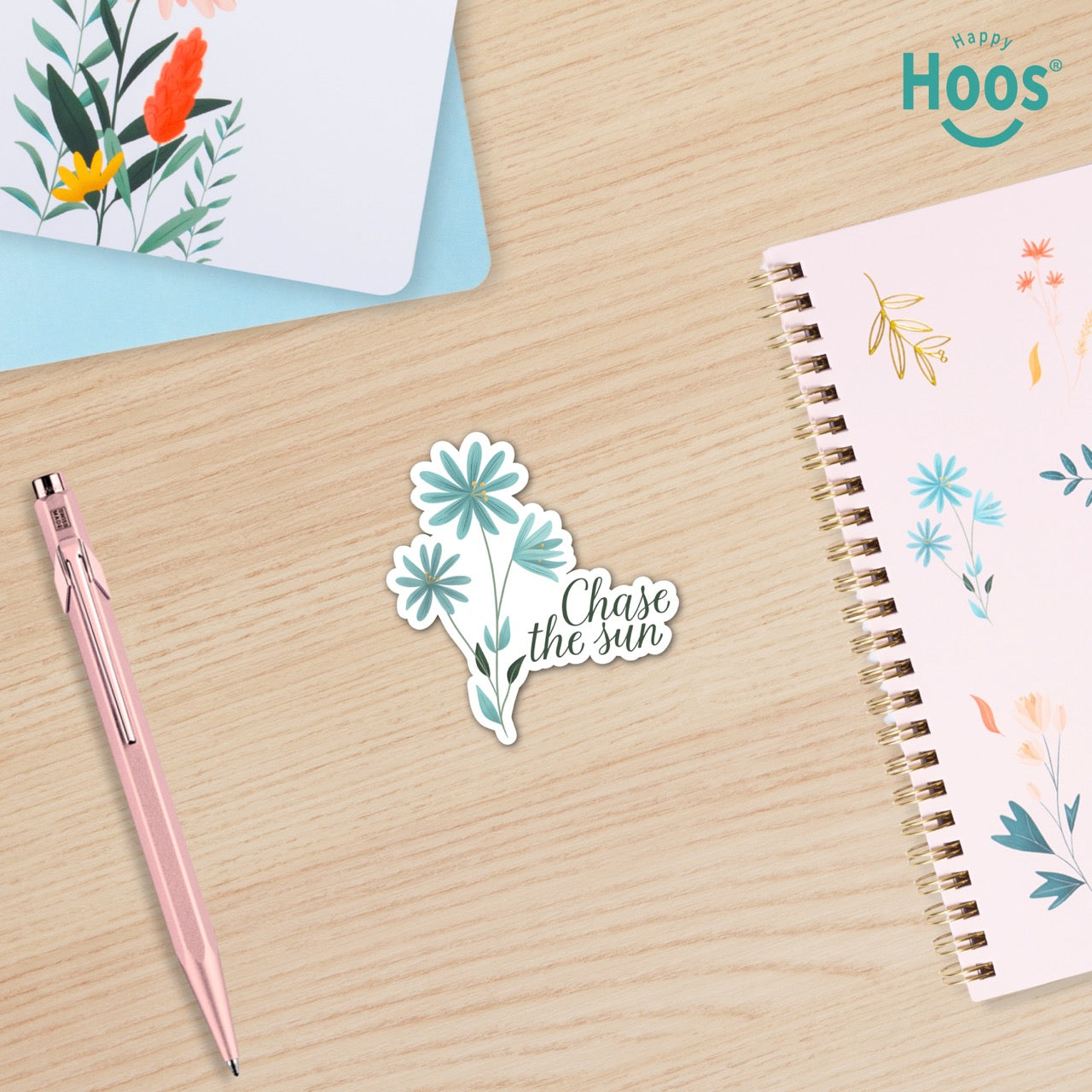 Sticker with floral design and 'Chase the sun' text on a wooden surface with a pen and notebook.