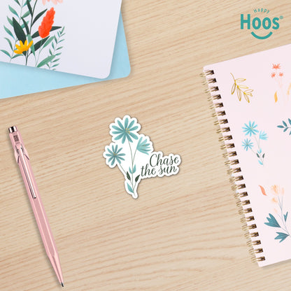 Sticker with floral design and 'Chase the sun' text on a wooden surface with a pen and notebook.