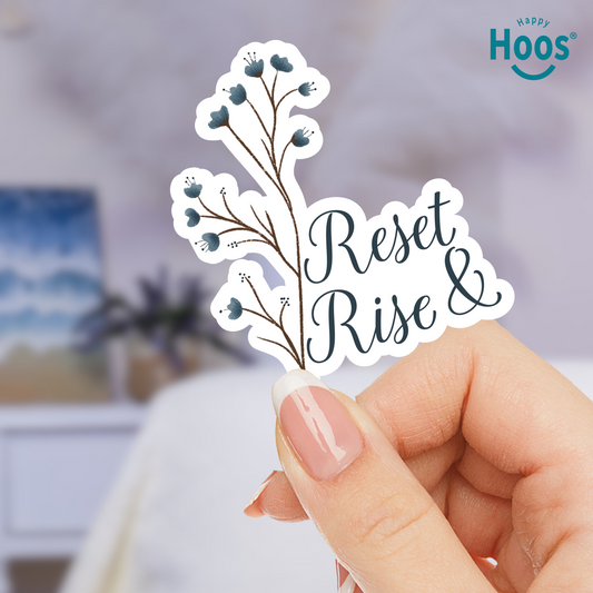 Hand holding a sticker with 'Reset & Rise' text and floral design, blurred indoor background