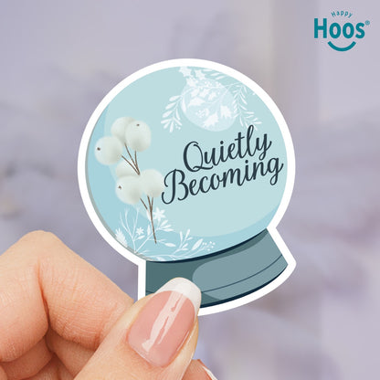 Round sticker with 'Quietly Becoming' text and floral design held by a hand, with 'Happy Hoos' logo in the corner.