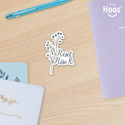 Sticker with 'Reset Rise &' text on a wooden surface with stationery items.