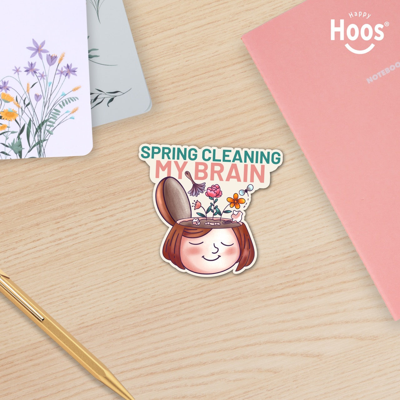 Spring Cleaning My Brain waterproof sticker on a wooden desk featuring a smiling face with flowers growing from an open head, made for self care, overthinkers, mental reset, planners, laptops, and water bottles.